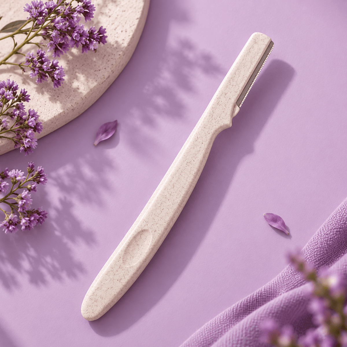 A long-handled facial razor with a textured off-white handle and a silver metal blade, styled on a lavender background with purple flowers and a stone dish.